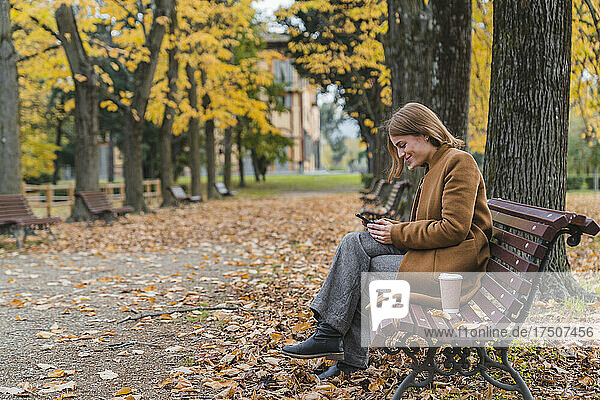 Lächelnde Frau benutzt Mobiltelefon auf Bank im Herbstpark