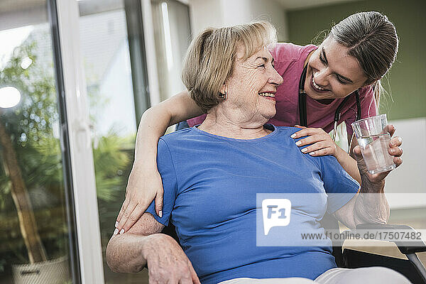 Smiling caregiver standing behind disabled woman in wheelchair at home
