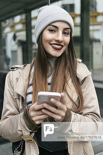Smiling woman with smart phone leaning on glass wall