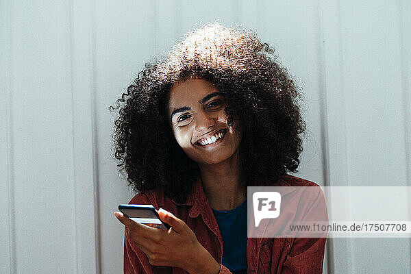 Lächelnde Frau mit Smartphone lehnt an weißer Wand