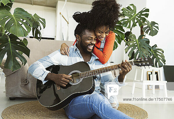 Junge Frau umarmt ihren Freund  der im Wohnzimmer Gitarre spielt