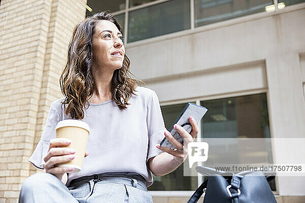 Contemplating businesswoman holding disposable cup and mobile phone