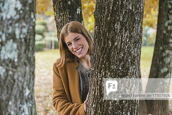 Happy young woman standing behind tree trunk