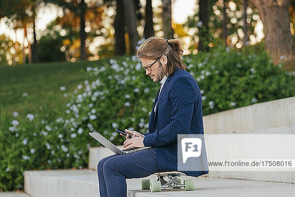 Businessman with laptop using mobile phone on skateboard