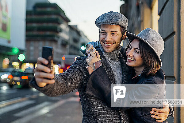 Happy young couple in hat taking selfie through smart phone in city