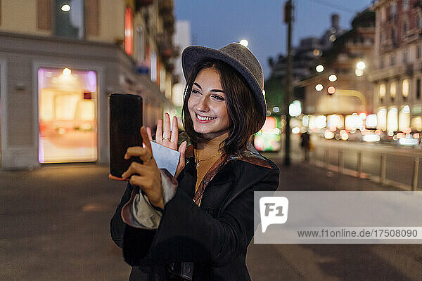 Glückliche junge Frau winkt während eines Videoanrufs über das Smartphone in der Stadt