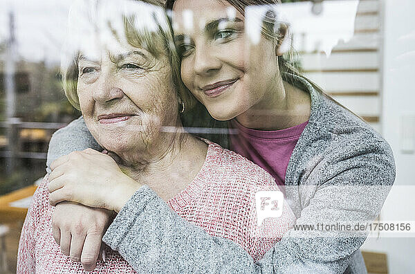 Young caregiver hugging senior woman looking out from window