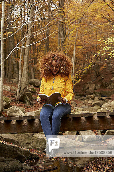 Smiling young woman reading book sitting on bridge in autumn forest