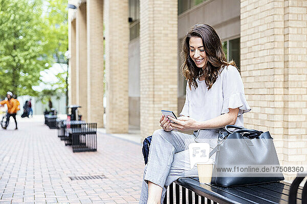 Smiling businesswoman using smart phone sitting on bench with bag and disposable cup
