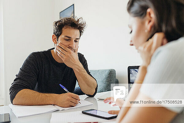 Young man with book and pen looking at businesswoman working on smart phone at home