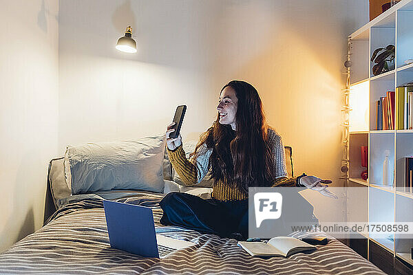 Woman doing video call though smart phone sitting with laptop on bed at home