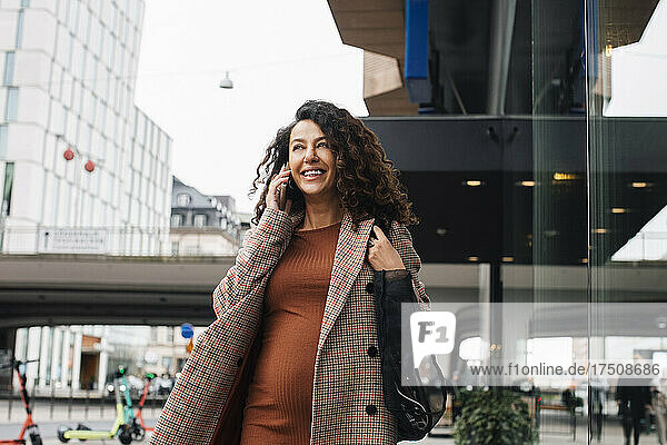 Lächelnde Geschäftsfrau  die beim Spaziergang in der Stadt mit ihrem Smartphone telefoniert