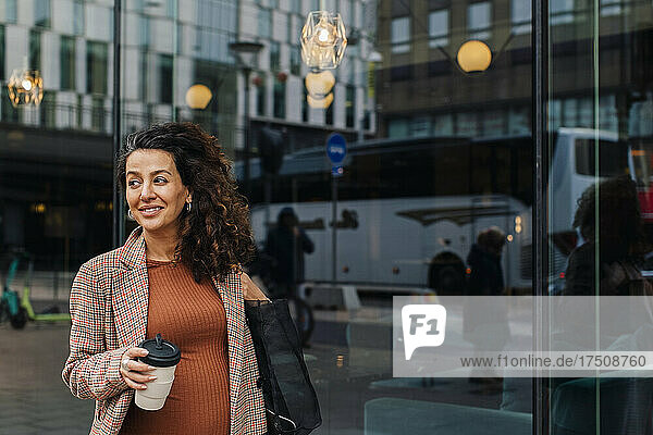 Lächelnde schwangere Geschäftsfrau mit Einweg-Kaffeebecher in der Stadt