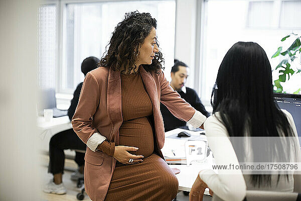 Pregnant businesswoman discussing with female colleague over computer in office