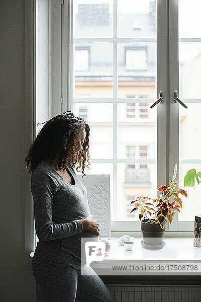 Schwangere Frau berührt ihren Bauch  während sie zu Hause am Fenster steht