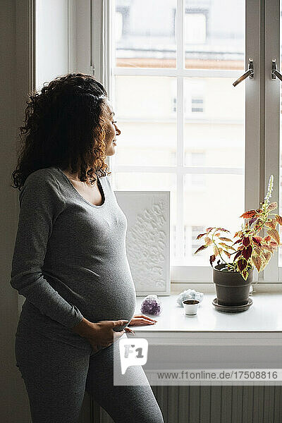 Schwangere Frau mit Hand auf dem Bauch schaut durch das Fenster zu Hause