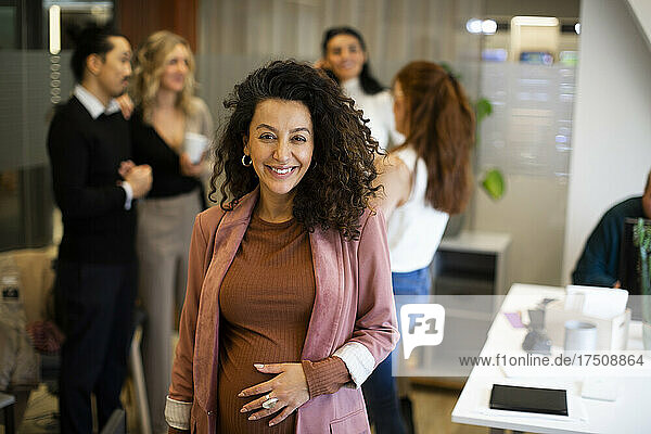 Porträt einer lächelnden schwangeren Geschäftsfrau mit Kollegen im Hintergrund im Büro