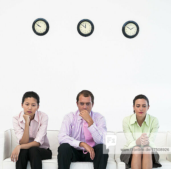 Drei Geschäftsleute sitzen auf einer Couch unter einer Uhr und wirken nervös.