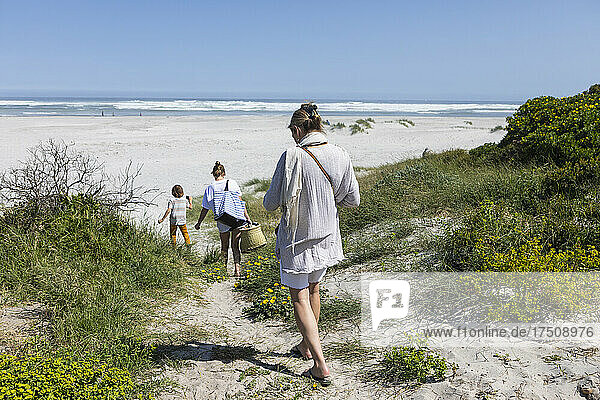 A family walking through the sand dunes towards the ocean with baskets and bags.