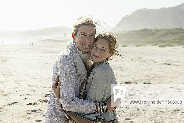 Erwachsene Frau und ihre Tochter im Teenageralter umarmen sich  sie stehen an einem windgepeitschten Strand