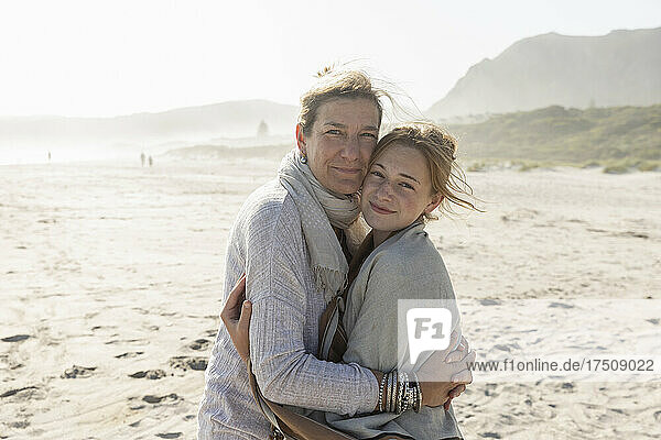 Erwachsene Frau und ihre Tochter im Teenageralter umarmen sich  sie stehen an einem windgepeitschten Strand