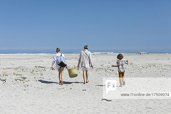 Mutter und Tochter im Teenageralter gehen an einem Sandstrand spazieren und tragen einen Korb  ein Junge folgt.
