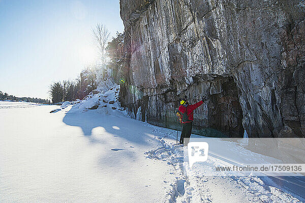 A man in a red jacket standing in front of an ice cliff looking up  ice climbing.