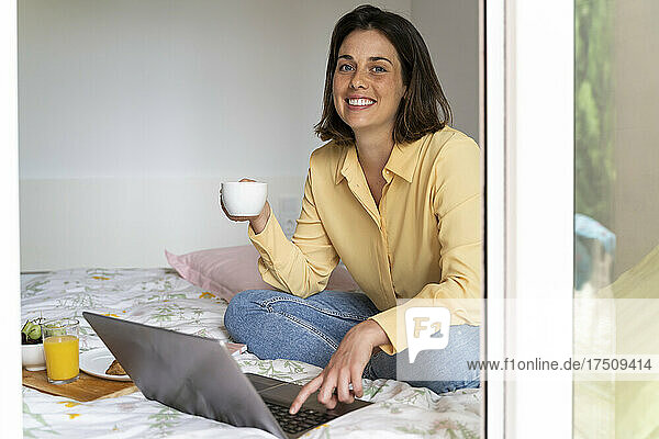 Smiling woman having coffee while using laptop in bedroom