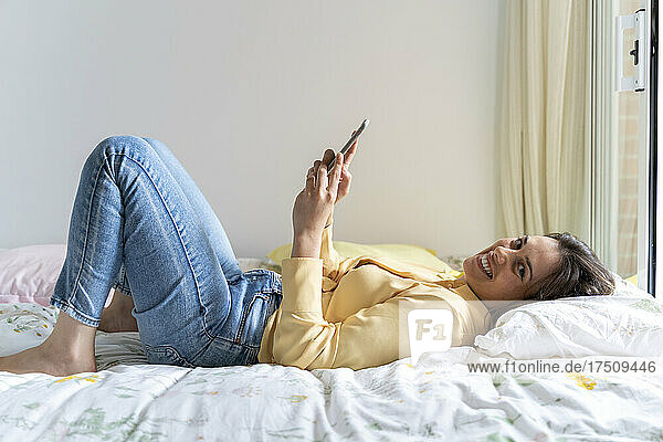 Smiling woman using mobile phone while lying in bedroom