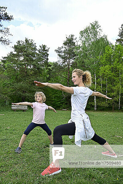Mother with daughter practicing warrior 2 pose on grassy land in forest