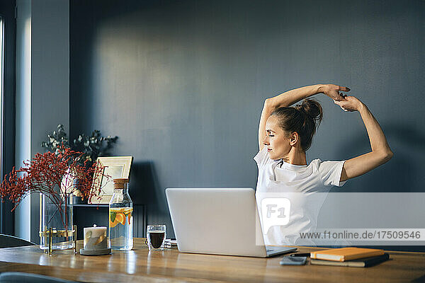 Tired businesswoman stretching arms while sitting at desk in home office