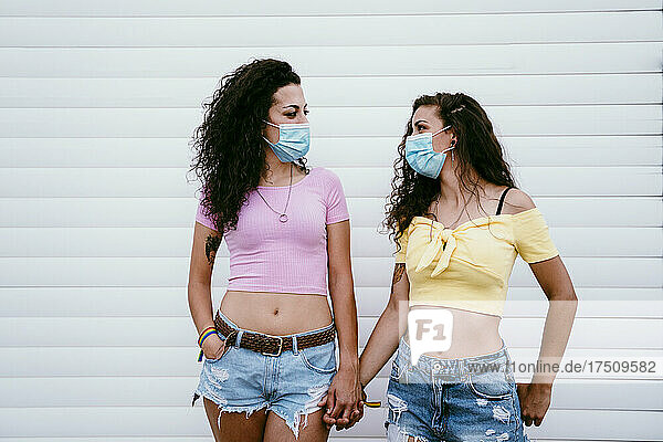 Lesbian couple wearing masks holding hands while standing against wall in city