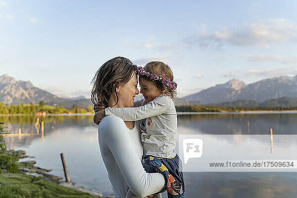 Mother carrying cheerful daughter while standing by lake against sky during sunset