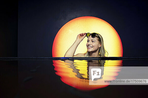 Lächelnde junge Frau mit Sonnenbrille schwimmt im Pool vor dem Hintergrund des Sonnenuntergangs