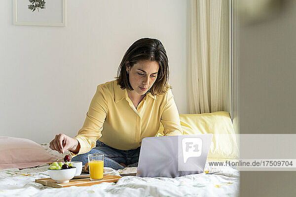 Woman eating breakfast while using laptop in bedroom
