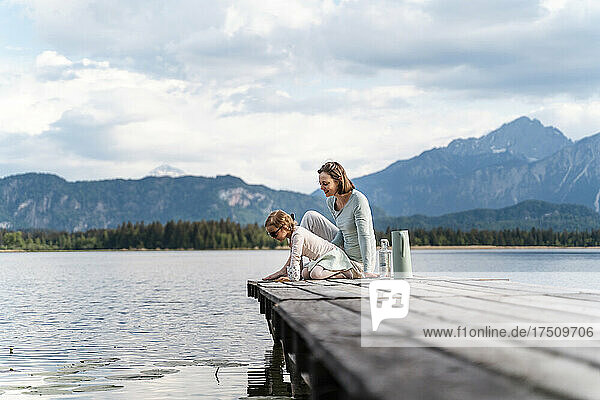 Mother with daughter sitting on jetty over lake against sky