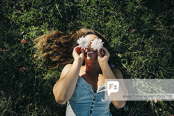 Young woman holding flowers over face while relaxing on grassy land in park