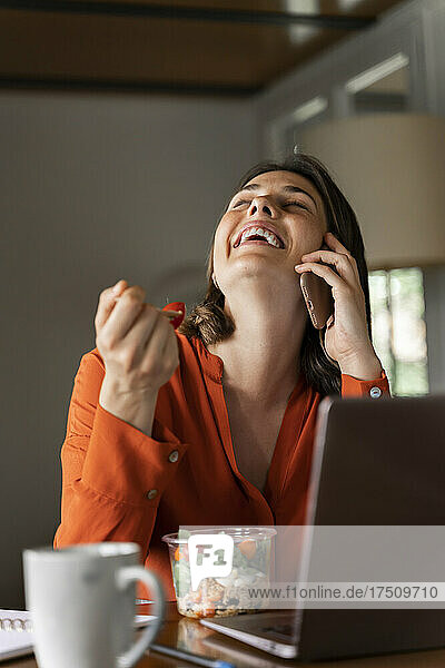 Happy businesswoman talking on mobile phone while eating salad at home