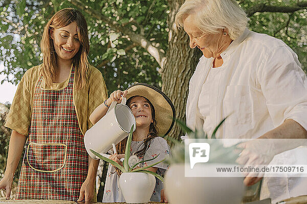 Smiling mother and grandmother looking at girl watering potted plant in yard