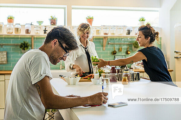Man making notes while women in background at kitchen island in cooking school