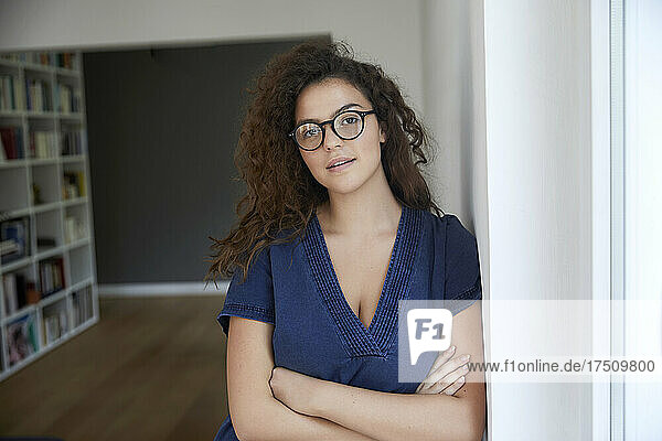 Beautiful woman wearing eyeglasses while leaning on wall with arms crossed at home
