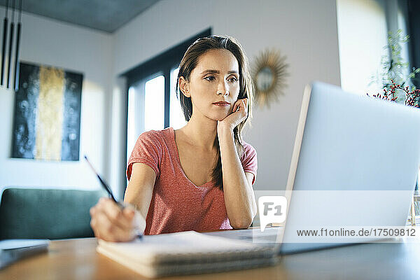 Serious female freelancer looking at laptop while writing in note pad on desk