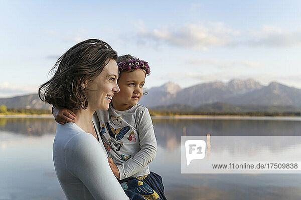 Smiling mother carrying cute daughter while standing against lake at sunset
