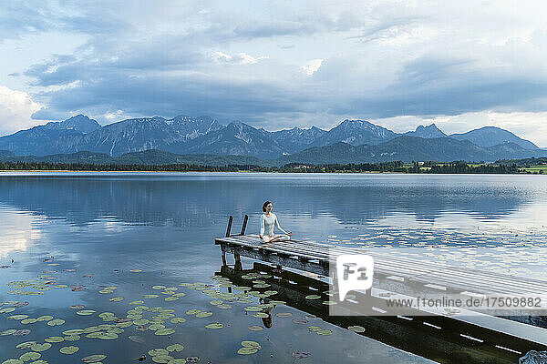 Frau meditiert  während sie auf einem Steg über dem See vor den Bergen sitzt