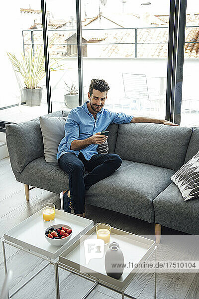 Mid adult man using smart phone while sitting on sofa in living room of modern penthouse