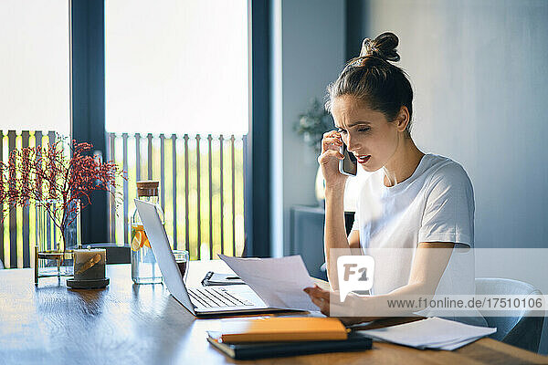Geschäftsfrau hält Dokument in der Hand und spricht über Smartphone am Schreibtisch im Heimbüro