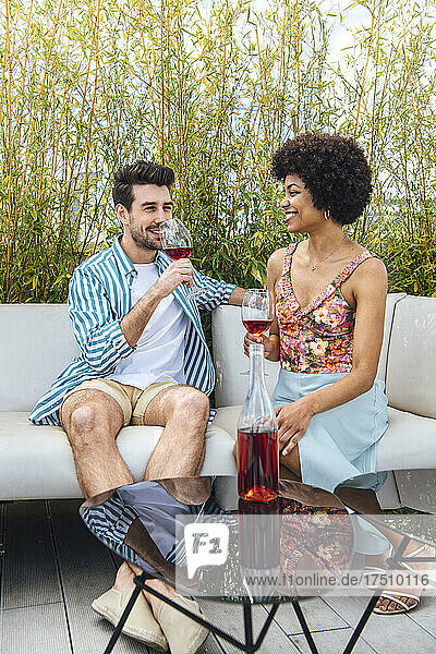 Happy multi-ethnic couple having red wine while sitting on sofa at penthouse patio