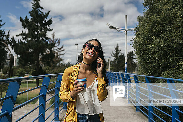 Fröhliche Frau mit Sonnenbrille spricht über Smartphone  während sie auf der Fußgängerbrücke im Park steht