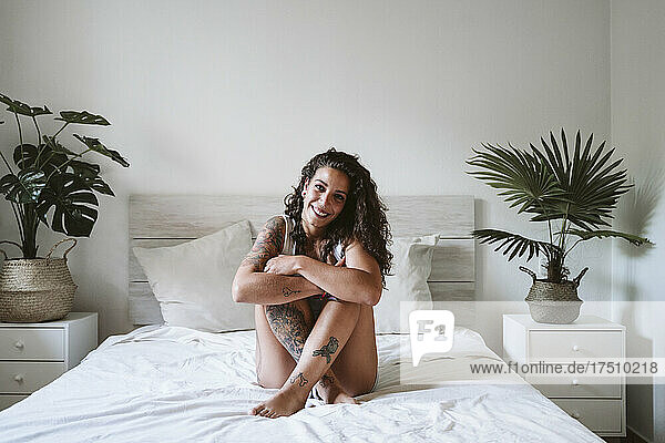 Lächelnde junge Frau umarmt Knie  während sie zu Hause auf dem Bett an der Wand sitzt