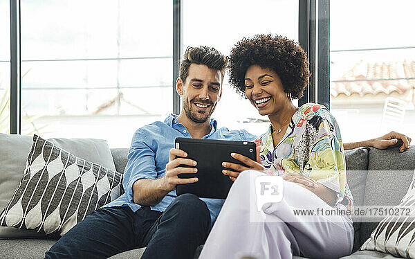 Happy multi-ethnic couple using digital tablet while sitting on sofa in penthouse
